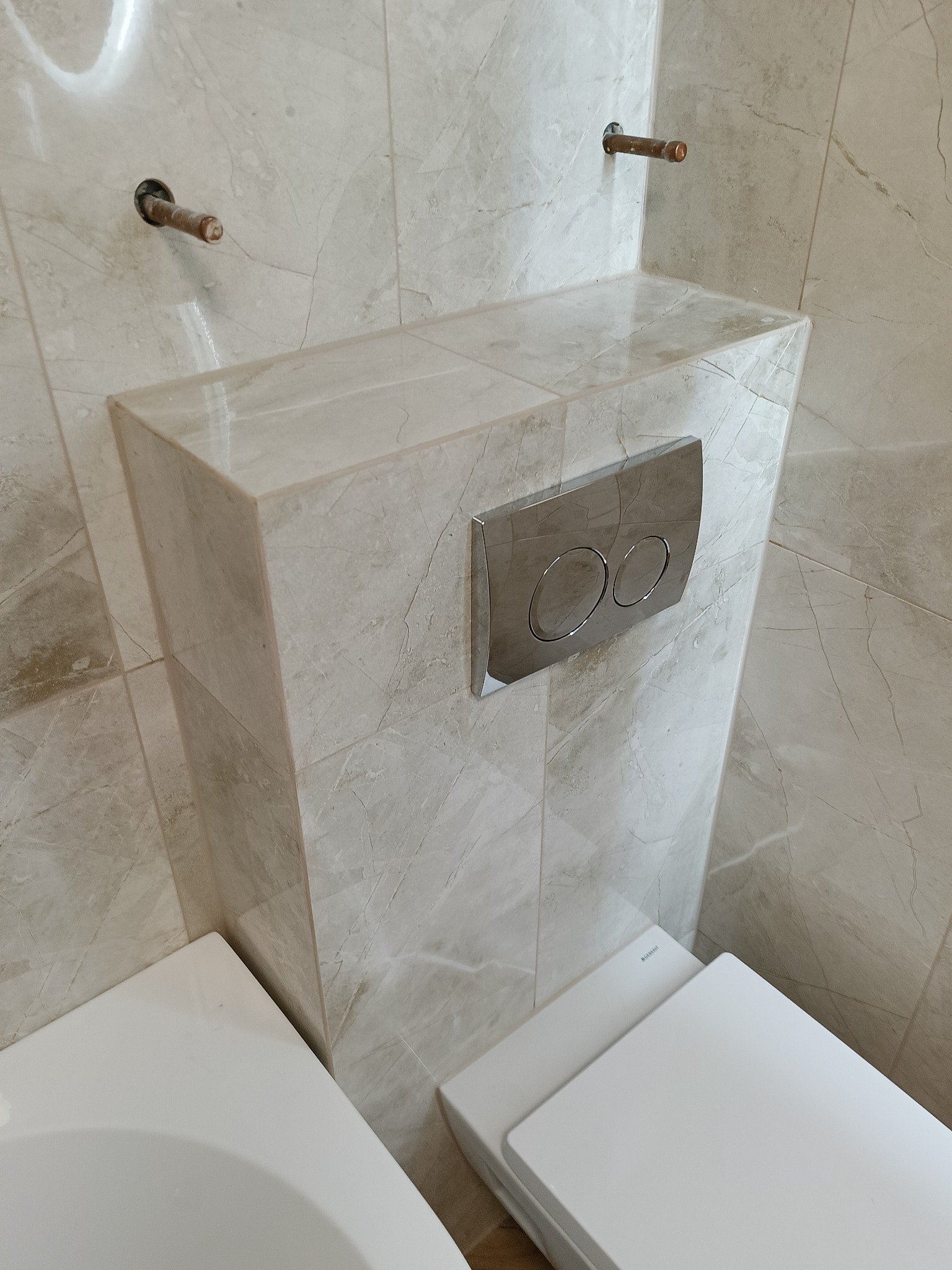 Wykończona obudowa stelaża WC z beżowych płytek imitujących marmur, z chromowanym przyciskiem spłukującym. Widoczne fragmenty toalety i rury doprowadzające wodę.