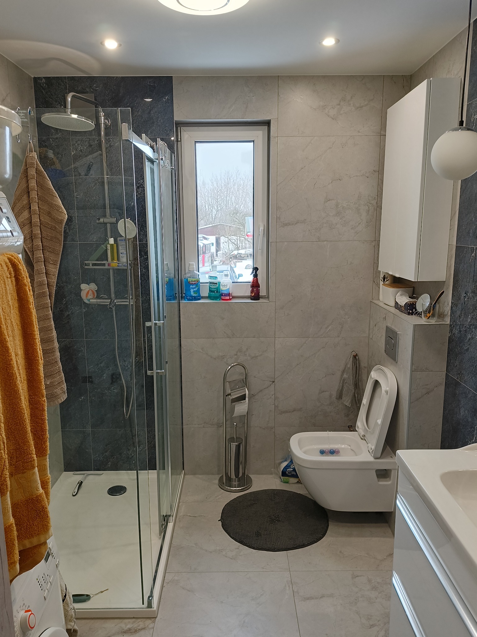 Jasna łazienka z kabiną prysznicową typu walk-in, szarą mozaiką i marmurowymi płytkami. Widoczne okno z widokiem na zimowy krajobraz. Nowoczesny design.