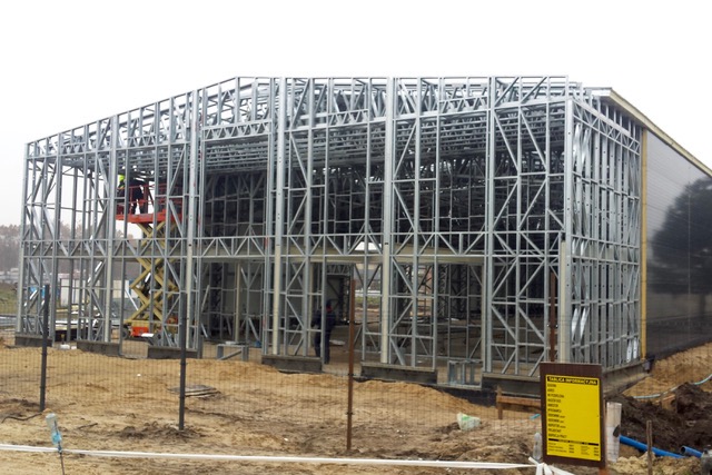 Wznoszenie stalowej konstrukcji hali: widoczny szkielet z metalowych profili, podnośnik koszowy z pracownikiem w górnej części, częściowo obłożona ściana, teren budowy w tle.