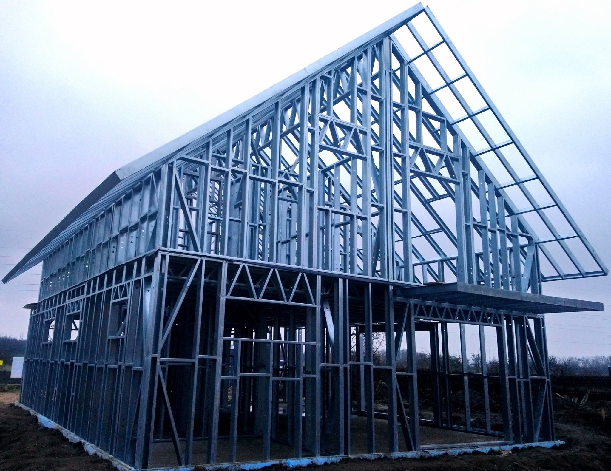 Metalowa konstrukcja szkieletowa domu jednorodzinnego w trakcie budowy, widoczne stalowe profile i elementy nośne, pochmurne tło.