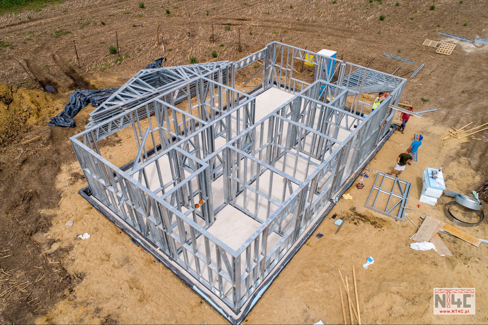 Widok z góry na stalowy szkielet domu w trakcie budowy, widoczni robotnicy w kamizelkach, przygotowane elementy konstrukcji, teren budowy z ziemią i resztkami materiałów.