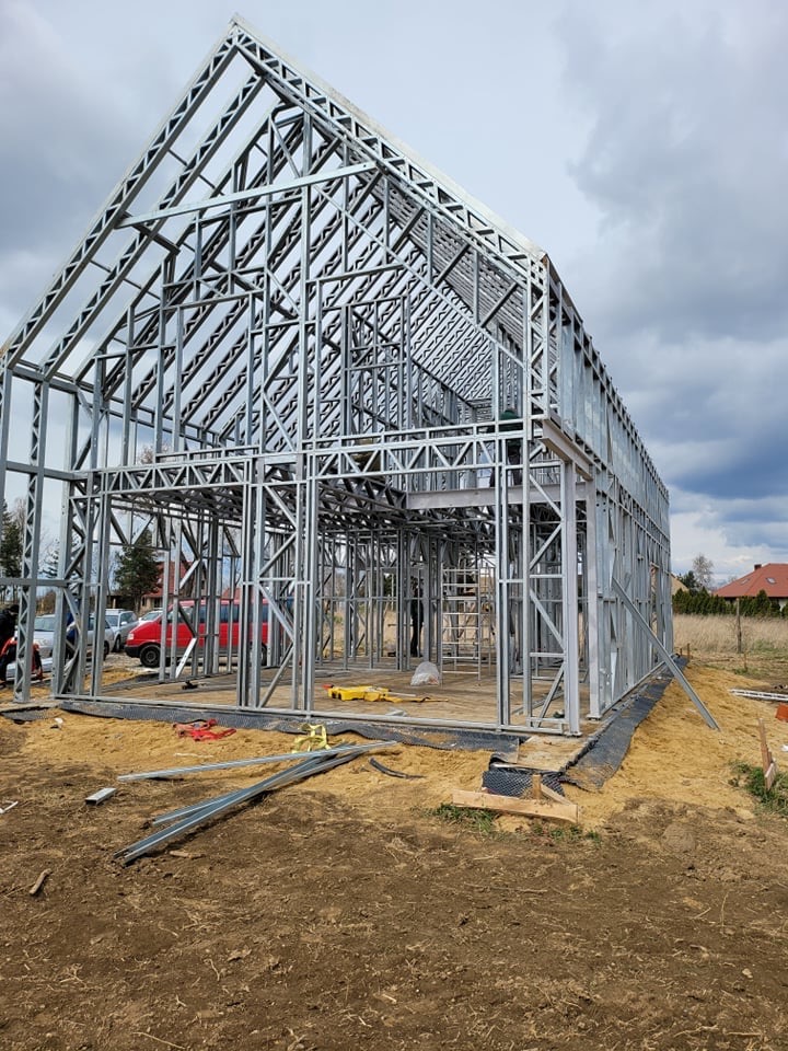 Konstrukcja domu ze stalowych profili, widoczne elementy szkieletu, narzędzia i materiały budowlane na placu budowy, pochmurne niebo w tle.