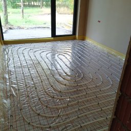 HYDRO-TECH WOJTASIK LESZEK - Białe rury ogrzewania podłogowego ułożone na folii z czerwoną siatką w surowym wnętrzu z dużym oknem wychodzącym na zieleń.