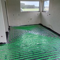 HYDRO-TECH WOJTASIK LESZEK - Zielone rury ogrzewania podłogowego ułożone na folii termoizolacyjnej w surowym wnętrzu z dwoma oknami, widok z góry. Instalacja przed zalaniem posadzką.