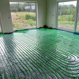 HYDRO-TECH WOJTASIK LESZEK - Zielone rury ogrzewania podłogowego ułożone na folii w jasnym pomieszczeniu z dużymi oknami, za którymi widać zieleń. Widok na instalację przed wylaniem posadzki.