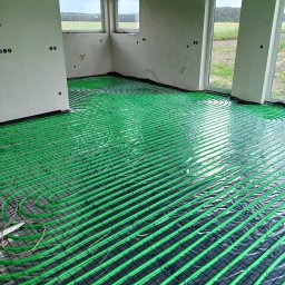 HYDRO-TECH WOJTASIK LESZEK - Jasnozielone rury ogrzewania podłogowego ułożone spiralnie na czarnej siatce w surowym wnętrzu z oknami. Widoczne niedokończone ściany i wystające kable.