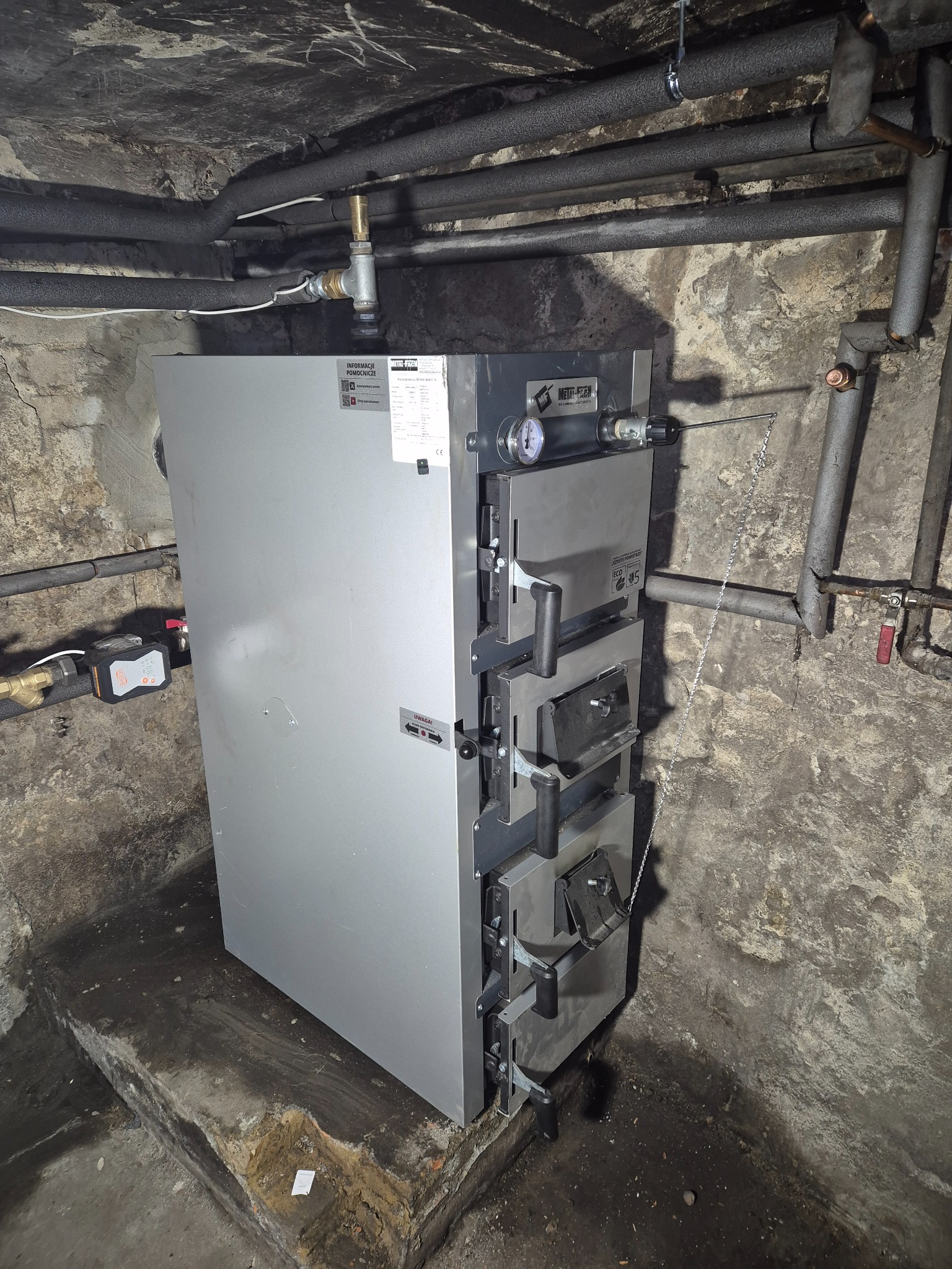 Nowoczesny piec CO z czterema drzwiczkami, zainstalowany w piwnicy z rurami grzewczymi i wilgotnymi ścianami z betonu.