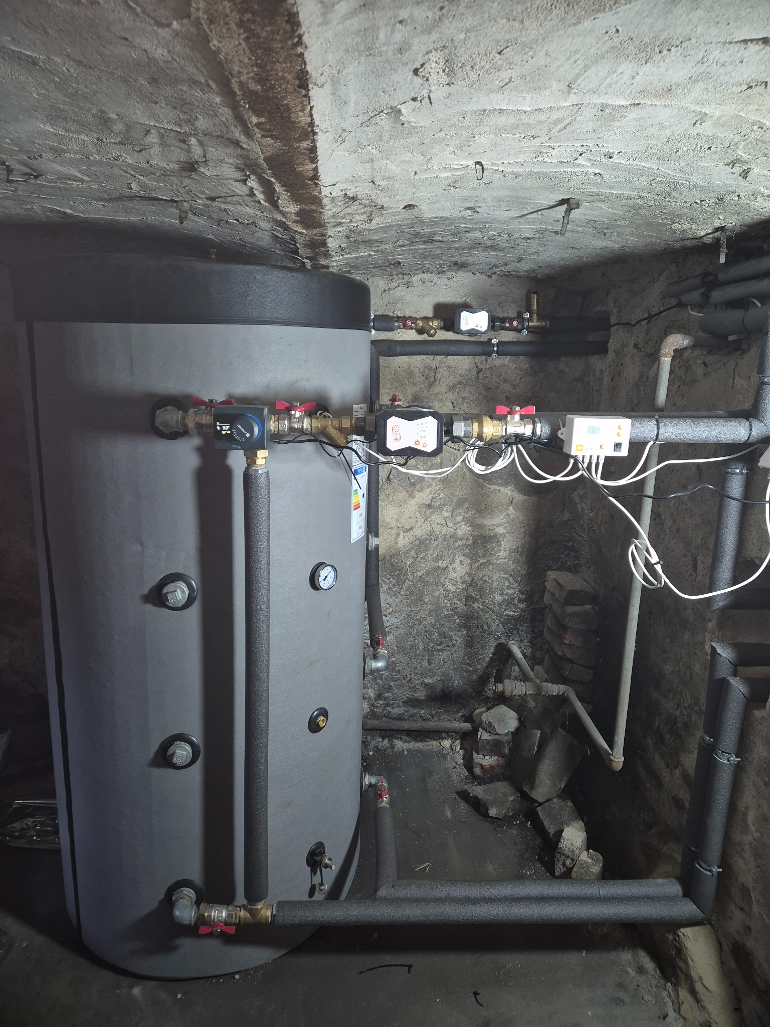 Szary, izolowany zbiornik na wodę z podłączonymi rurami i armaturą w piwnicy z surowym betonowym sufitem.