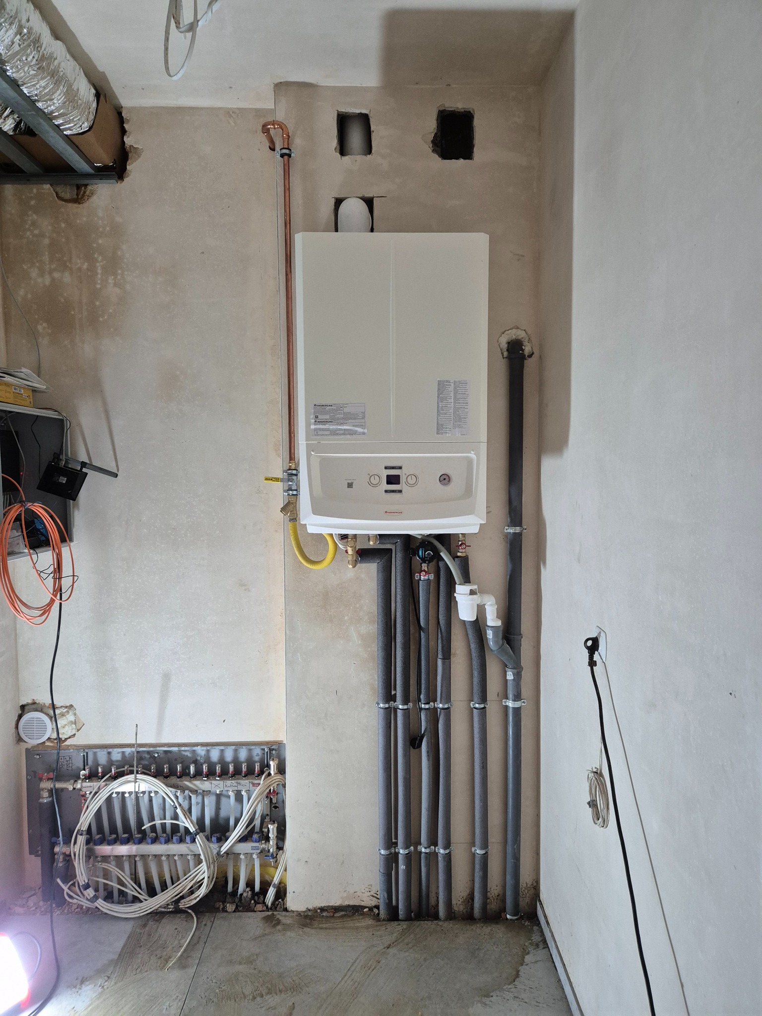 Biały piec centralnego ogrzewania marki Viessmann zamontowany na szarej, surowej ścianie, z widocznymi rurami instalacji grzewczej i rozdzielaczem ogrzewania podłogowego w dolnej części kadru.