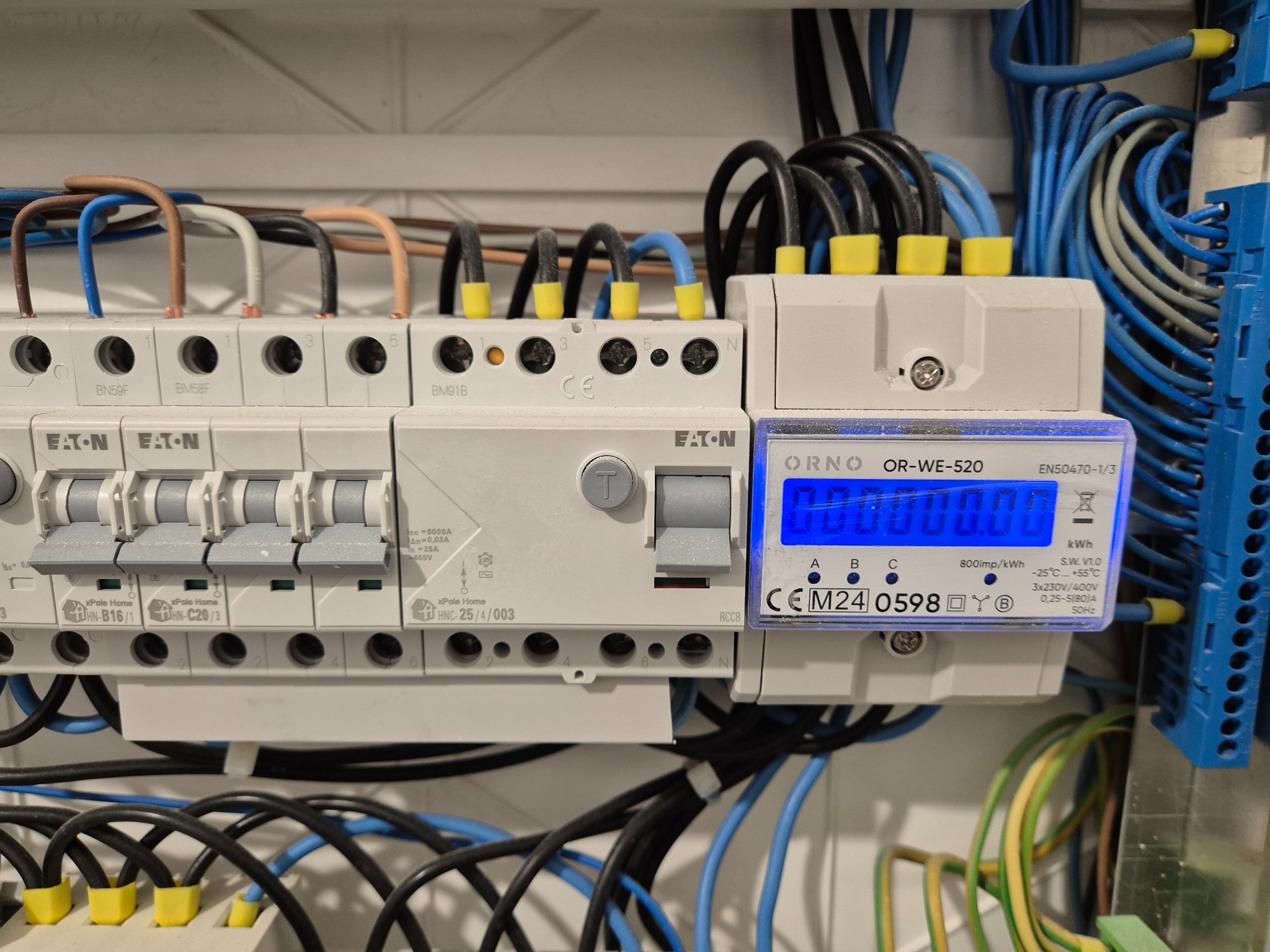 Szczegółowe ujęcie tablicy rozdzielczej z bezpiecznikami Eaton i licznikiem energii Orno, z widocznymi przewodami w różnych kolorach i oznaczeniami na elementach.