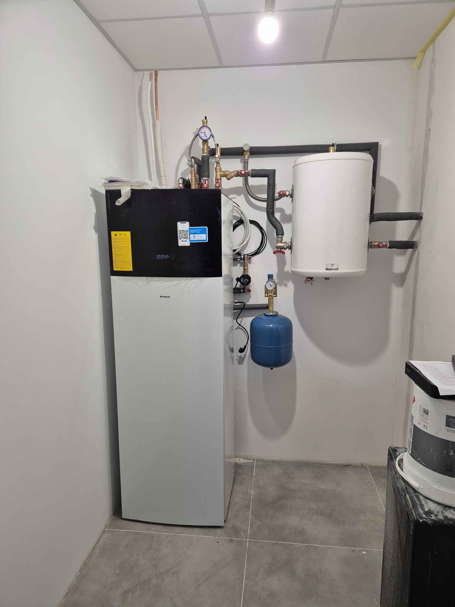 Nowoczesna pompa ciepła Daikin w kotłowni, z podłączonym zbiornikiem buforowym Galmet i naczyniem przeponowym, instalacja w trakcie podłączania, widoczne rury miedziane i izolowane rury grzewcze.