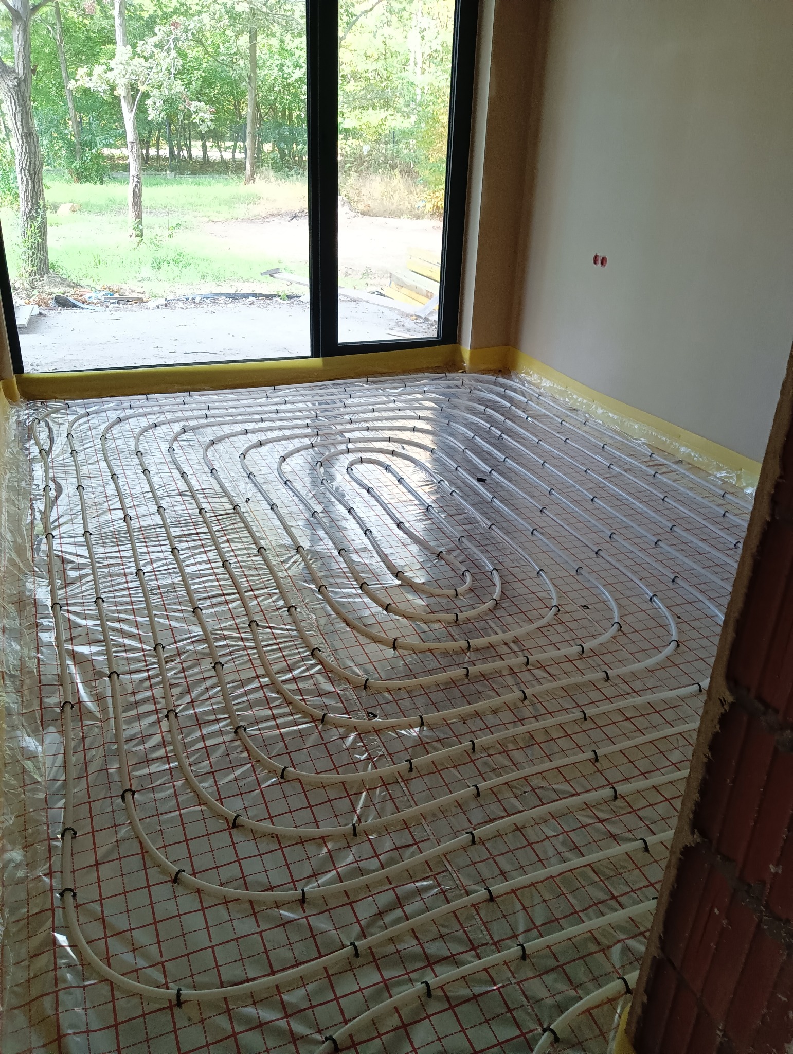 Białe rury ogrzewania podłogowego ułożone na folii z czerwoną siatką w surowym wnętrzu z dużym oknem wychodzącym na zieleń.