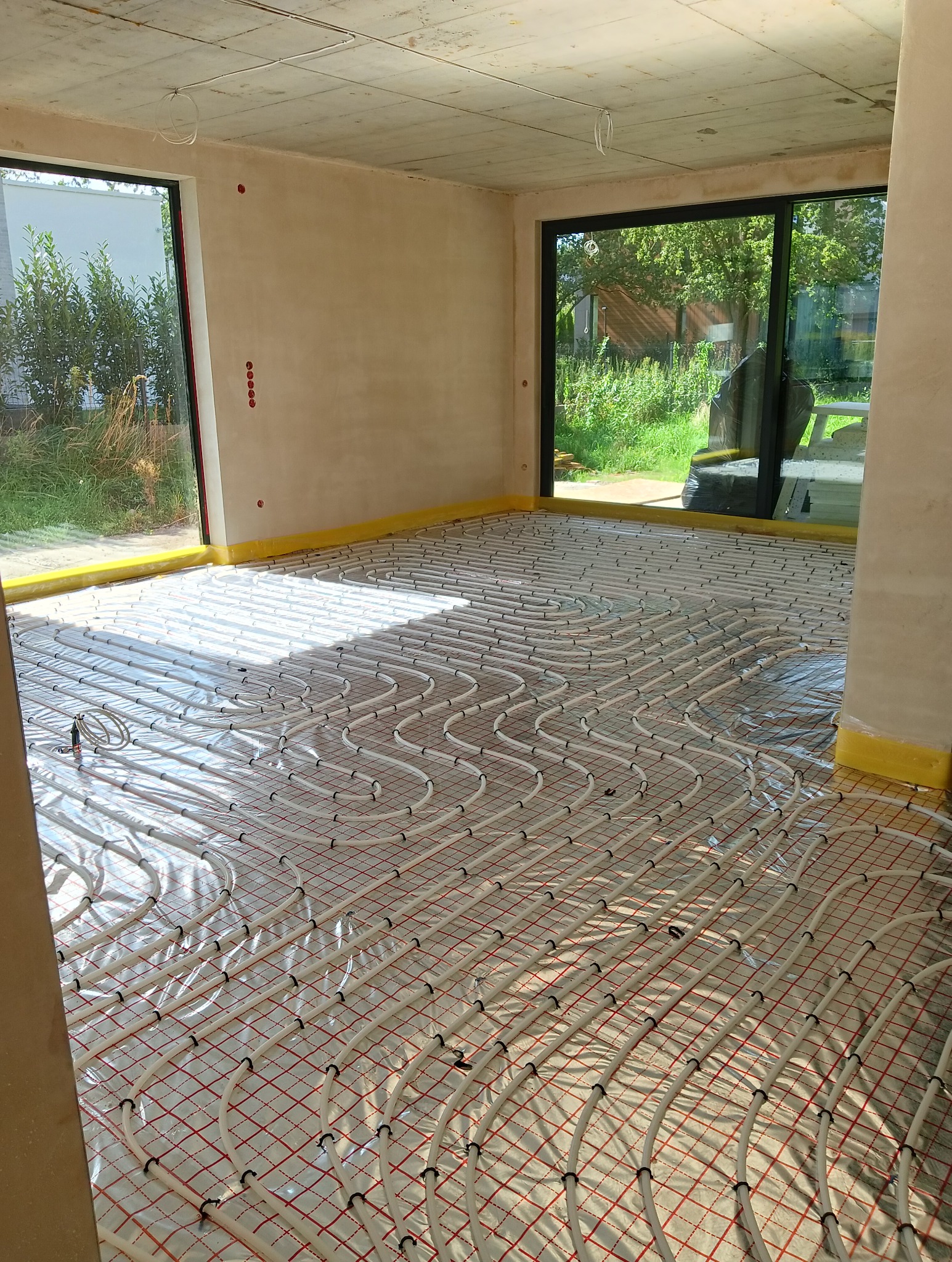 Rozłożona instalacja ogrzewania podłogowego z rurkami na folii izolacyjnej w surowym pomieszczeniu z dużymi oknami.