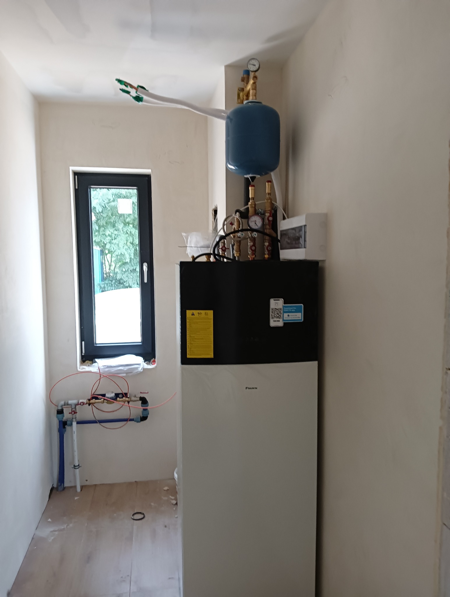 Biała pompa ciepła Daikin z niebieskim zbiornikiem wyrównawczym i instalacją hydrauliczną w pomieszczeniu w trakcie wykańczania, widoczne rury i okno z widokiem na zieleń.