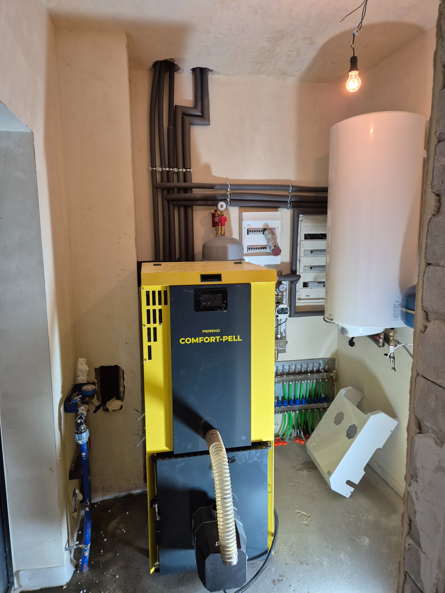 Żółto-czarny piec CO Pereko Comfort-Pell w kotłowni, z widoczną instalacją grzewczą, rurami w izolacji i białym zasobnikiem ciepłej wody. Surowy stan ścian.