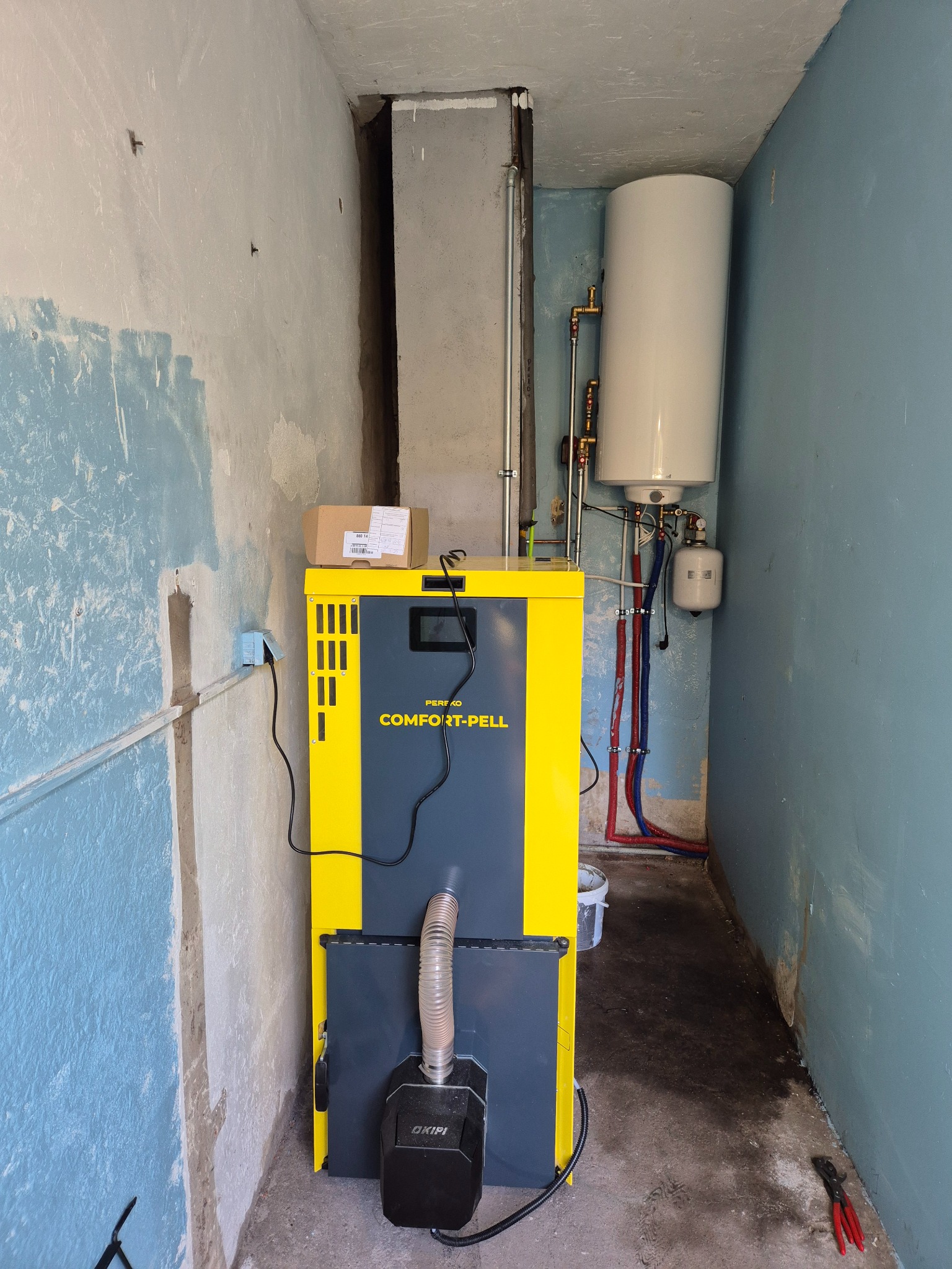 Żółto-szary piec CO Comfort-Pell z podłączonymi rurami i zbiornikiem na wodę w pomieszczeniu z surowymi ścianami, widoczne narzędzia i karton na piecu.
