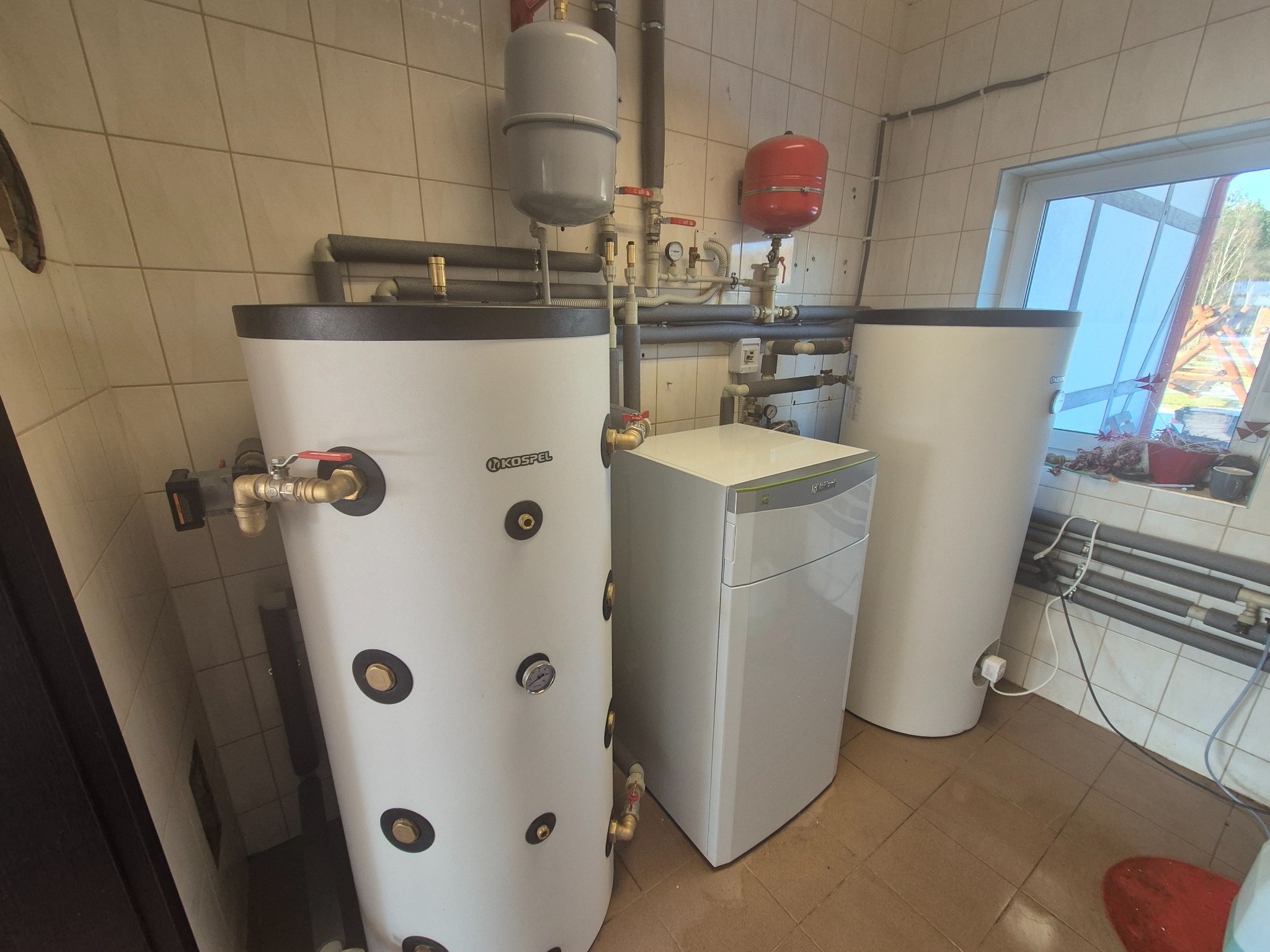 Kotłownia z białym zasobnikiem Kospel, piecem gazowym Vaillant i czerwonym naczyniem przeponowym. Rury grzewcze z izolacją widoczne na ścianie w płytkach.