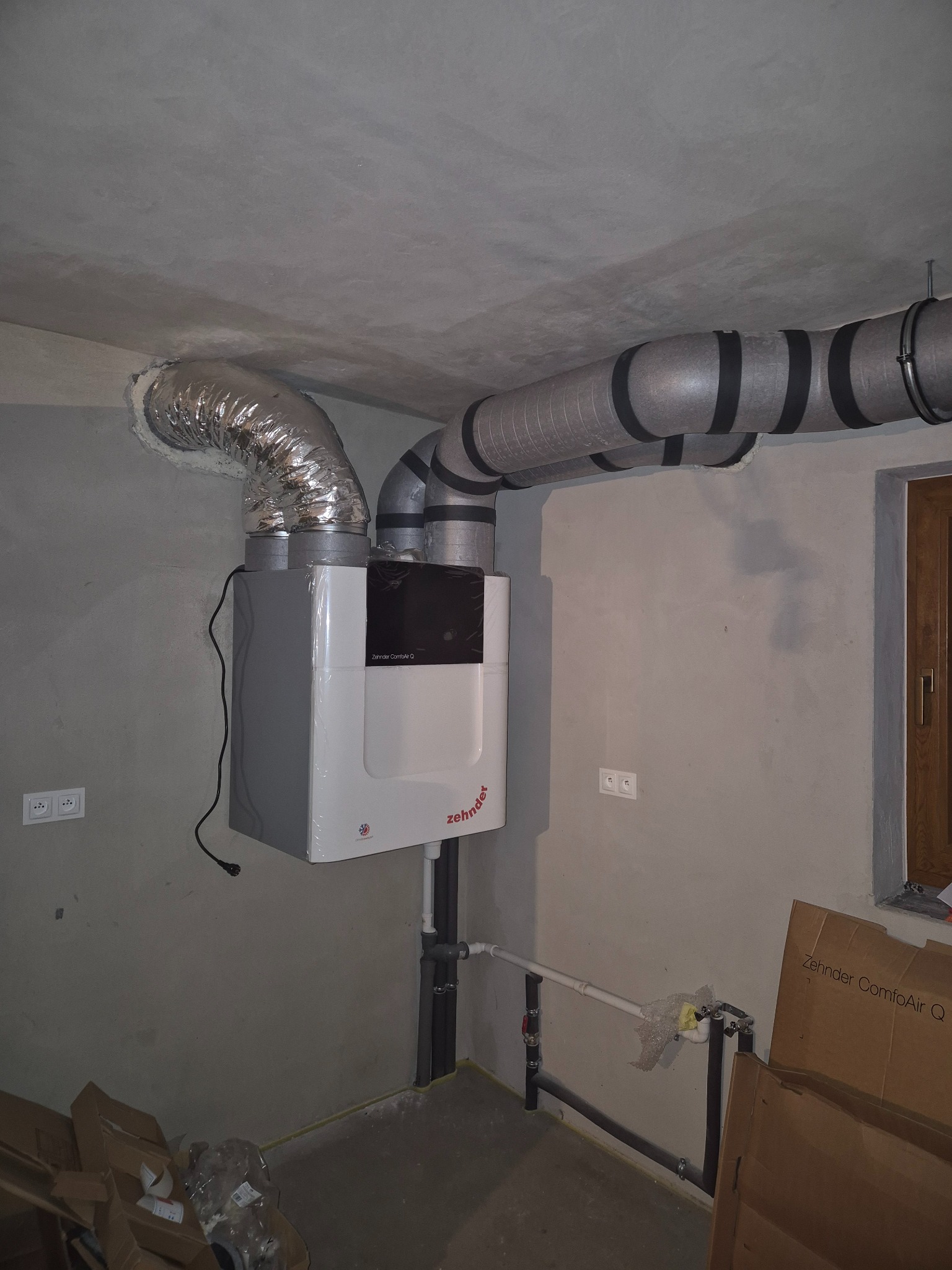 Centrala wentylacyjna Zehnder ComfoAir Q z podłączonymi przewodami wentylacyjnymi w surowym wnętrzu. Widoczne rury i kable, surowy beton na suficie.