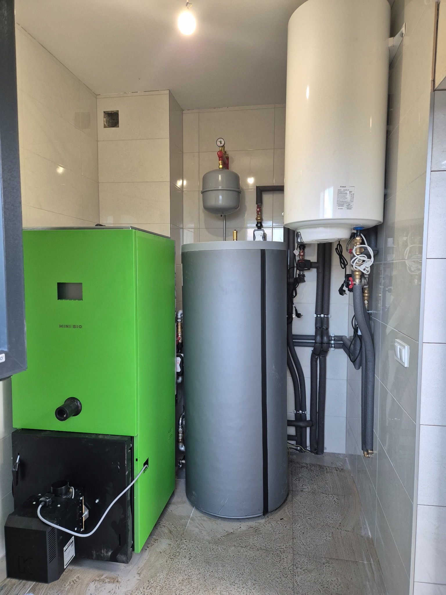 Zestaw urządzeń grzewczych w kotłowni: zielony piec Mini Bio, szary zbiornik buforowy, biały bojler wiszący. Widoczne rury instalacji CO i CWU. Tło z płytek ceramicznych.