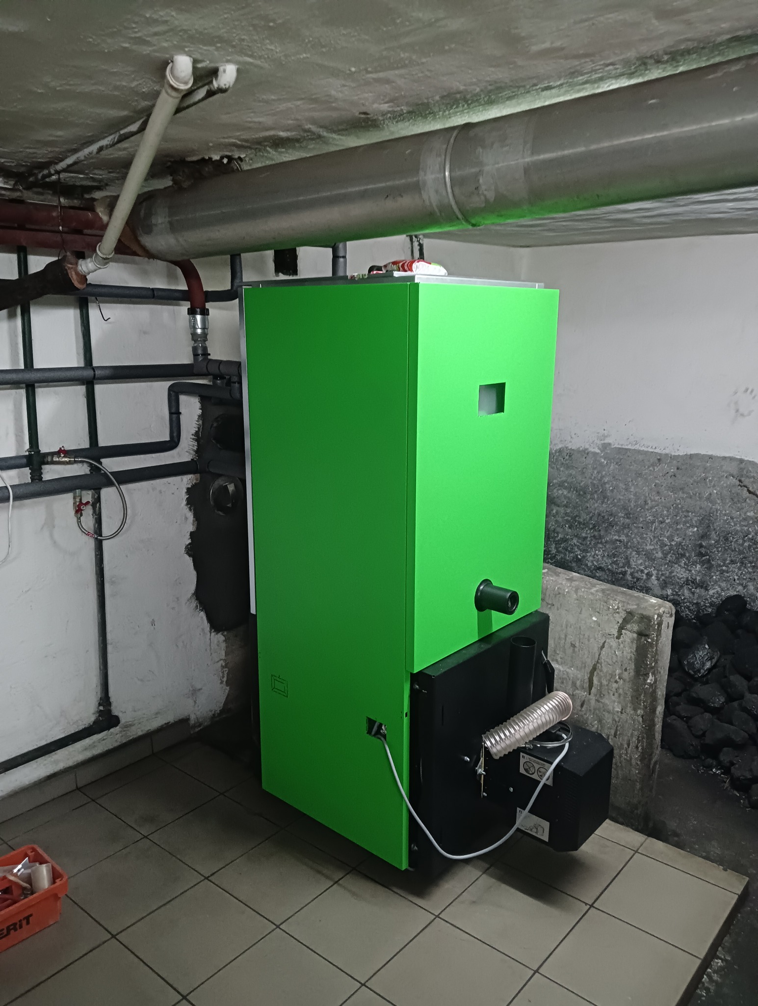 Zielony piec CO w kotłowni z instalacją i zapasem węgla. Widoczne rury, podłączenia. Narożnik z węglem dodaje realizmu.