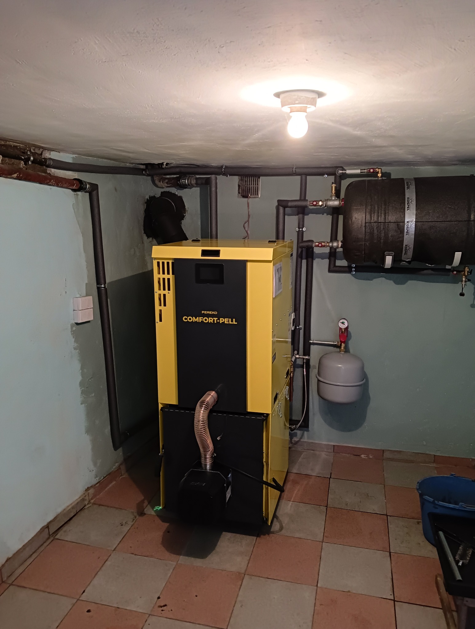 Żółto-czarny piec Comfort-Pell Pereko w kotłowni z instalacją CO, widoczne rury i zbiornik wyrównawczy. Nierówne ściany i płytki podłogowe.