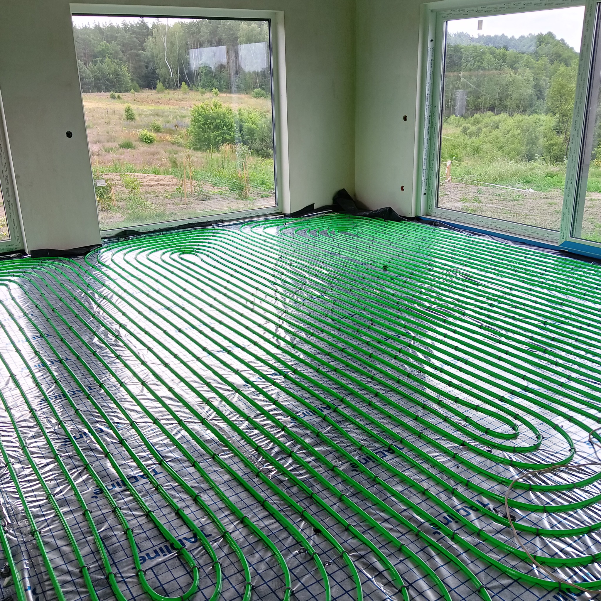 Zielone rury ogrzewania podłogowego ułożone na folii w jasnym pomieszczeniu z dużymi oknami, za którymi widać zieleń. Widok na instalację przed wylaniem posadzki.