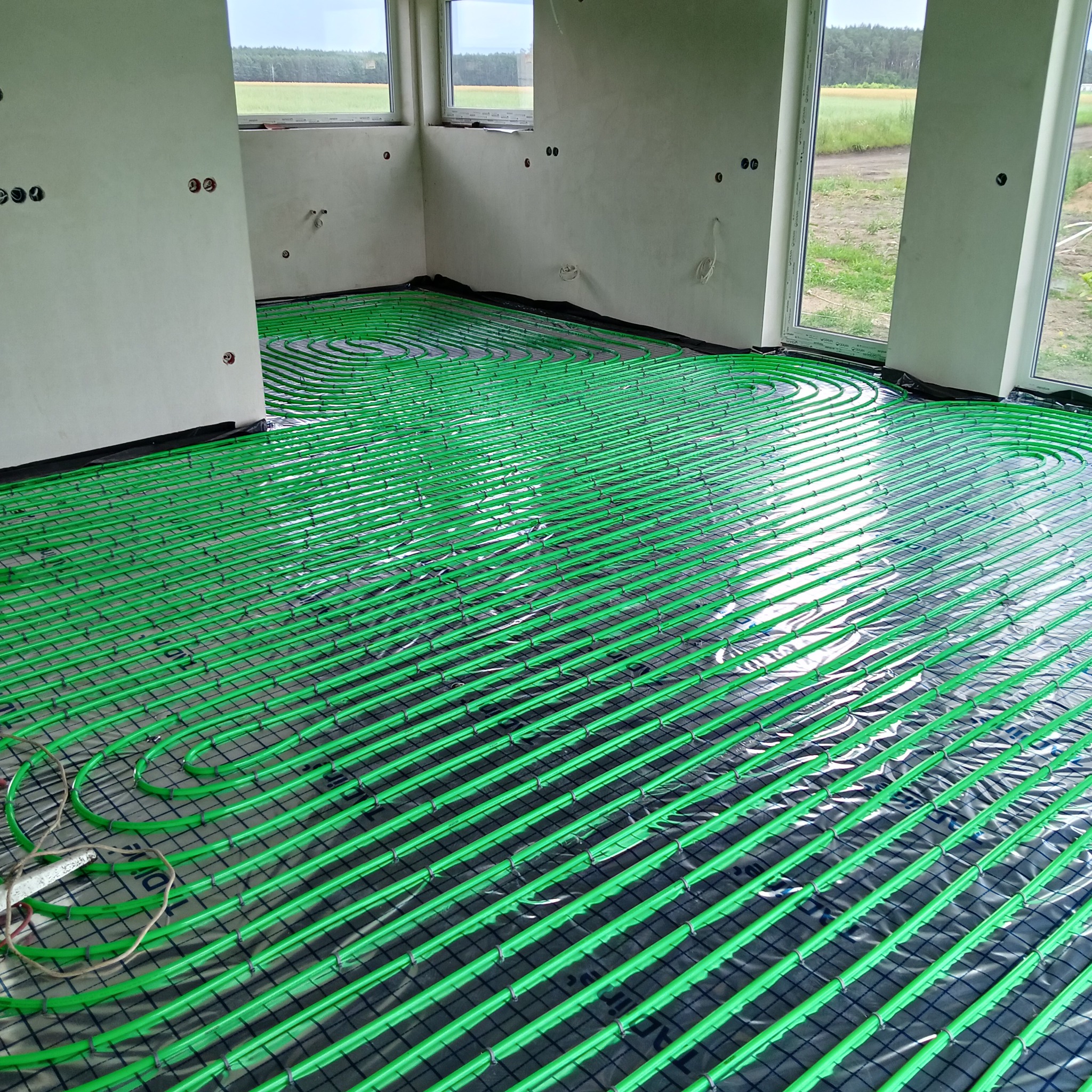 Jasnozielone rury ogrzewania podłogowego ułożone spiralnie na czarnej siatce w surowym wnętrzu z oknami. Widoczne niedokończone ściany i wystające kable.