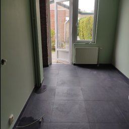 Polstal-Artur Bernekier - Pomalowane na zielono wnętrze pokoju z czarną podłogą z płytek, grzejnikiem pod oknem i otwartymi drzwiami balkonowymi z widokiem na ogród.