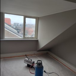 Polstal-Artur Bernekier - Pokój na poddaszu z oknem, świeżo pomalowanymi ścianami w odcieniach szarości i białym sufitem. Na podłodze leży folia ochronna, a obok okna stoi nagrzewnica gazowa z podłączonym przewodem i butlą...