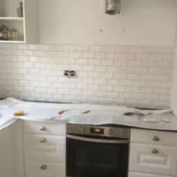 Układanie białych, prostokątnych płytek na ścianie w kuchni, widoczne gniazdka elektryczne bez osprzętu, zabezpieczony blat z narzędziami glazurniczymi i fragmentem płytki.