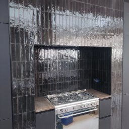 Artur Bernekier - Nowoczesna kuchnia z ciemnymi, błyszczącymi, pionowymi płytkami o nieregularnej fakturze, tworzącymi wnękę nad wolnostojącą kuchenką gazową, obok matowych szafek.