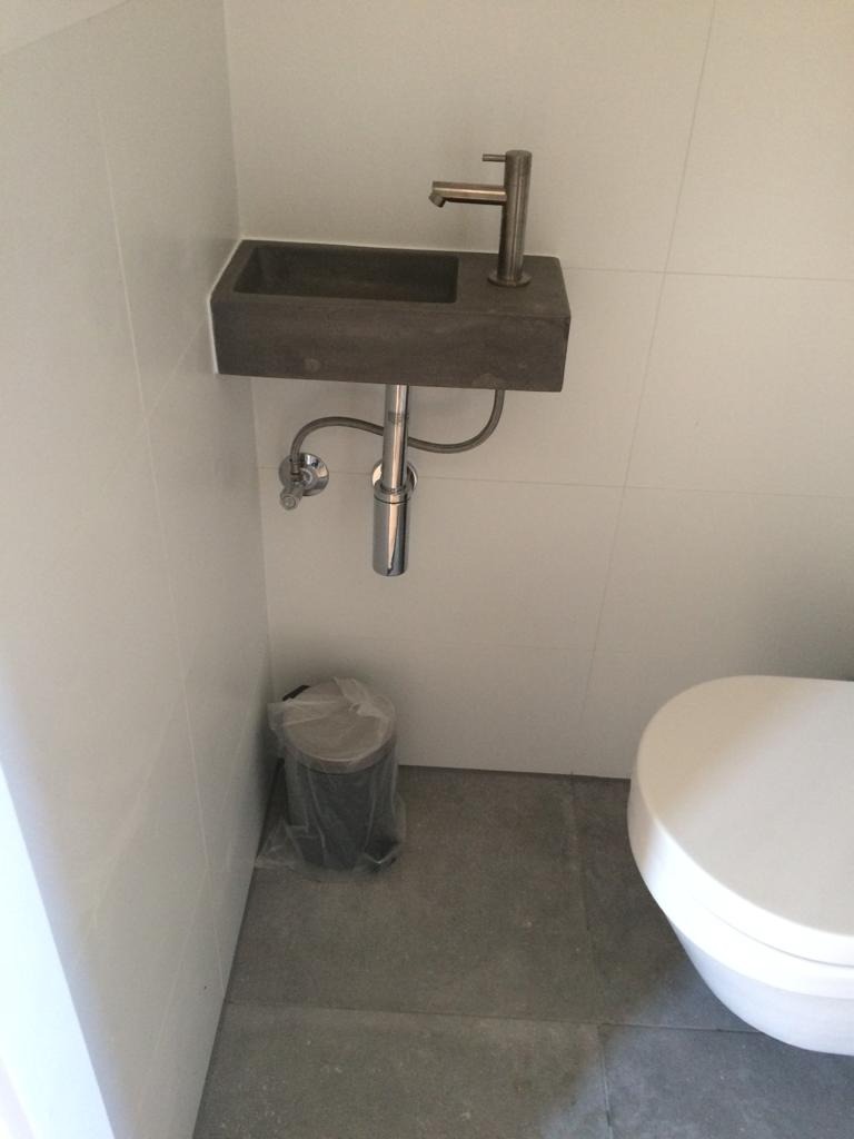 Mała, szara umywalka z chromowaną baterią zamontowana na białej ścianie w łazience z szarą podłogą, obok toaleta i kosz na śmieci w folii.