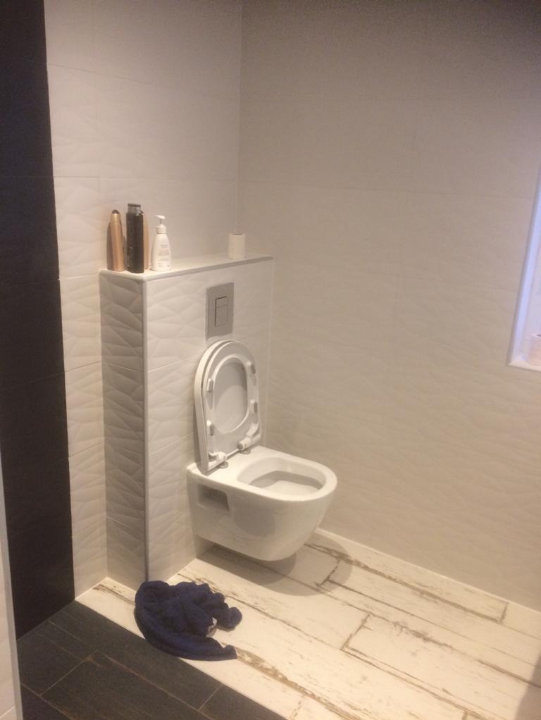 Wisząca toaleta z otwartą klapą, zabudowana w ścianie wyłożonej płytkami z subtelnym wzorem, na podłodze imitacja drewna, obok leży niebieski ręcznik.