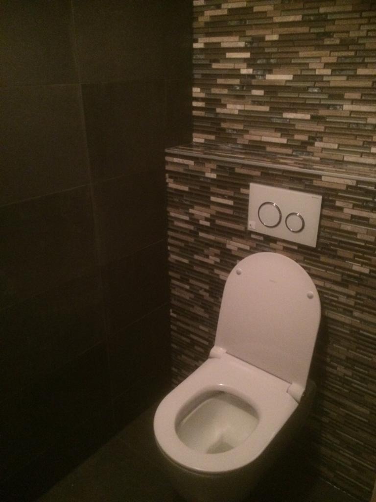 Toaleta z otwartą klapą w łazience wykończonej ciemnymi płytkami i mozaiką z beżowych i brązowych pasków, widoczny panel spłuczki z dwoma przyciskami.