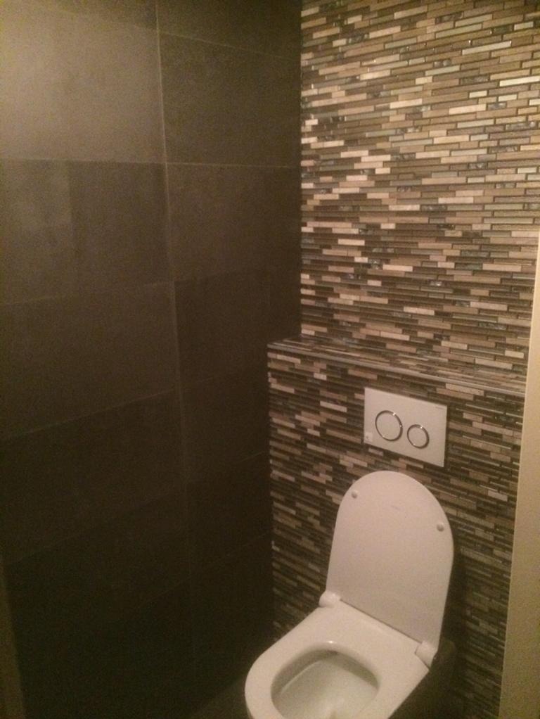 Nowoczesna toaleta podwieszana z białym przyciskiem spłukującym na ścianie wyłożonej mozaiką z drobnych, poziomych płytek w odcieniach beżu i brązu, kontrastująca z ciemnymi, gładkimi płytkami po...