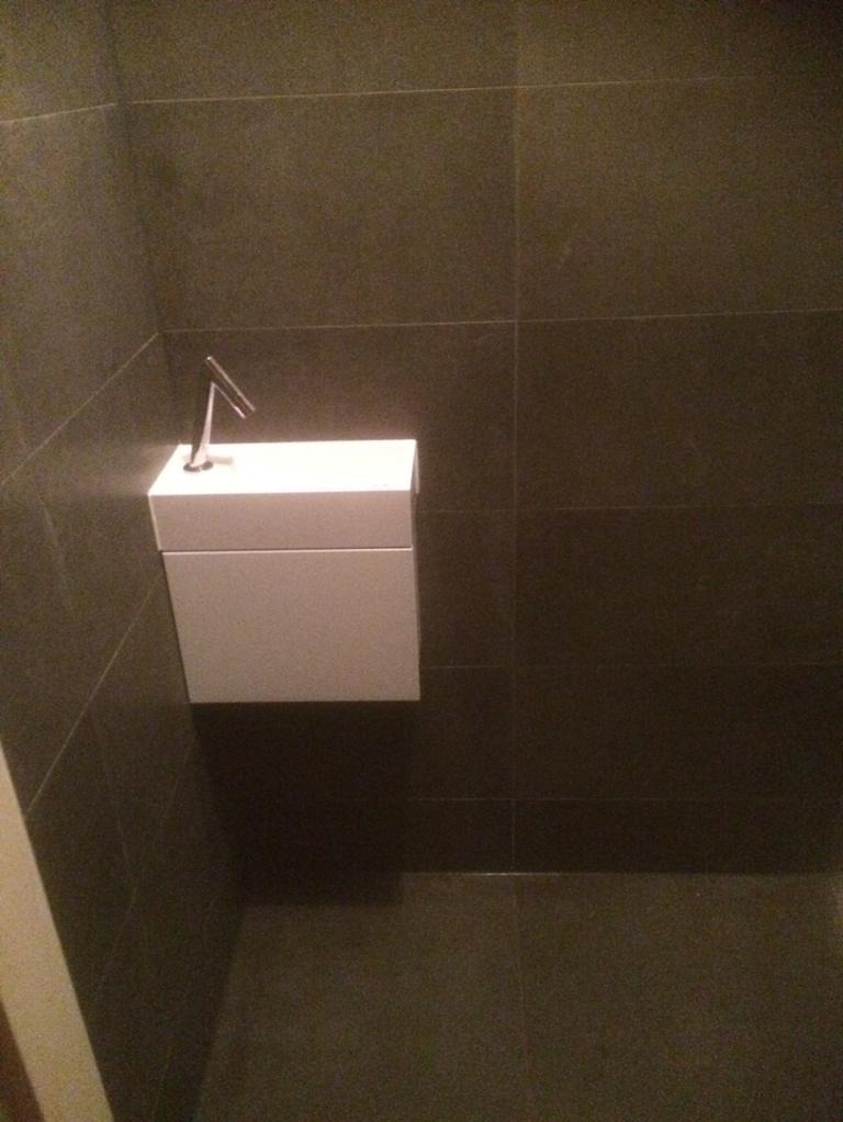 Minimalistyczna, wisząca umywalka o kwadratowym kształcie z chromowaną baterią, na tle ciemnych, grafitowych płytek ściennych i podłogowych.