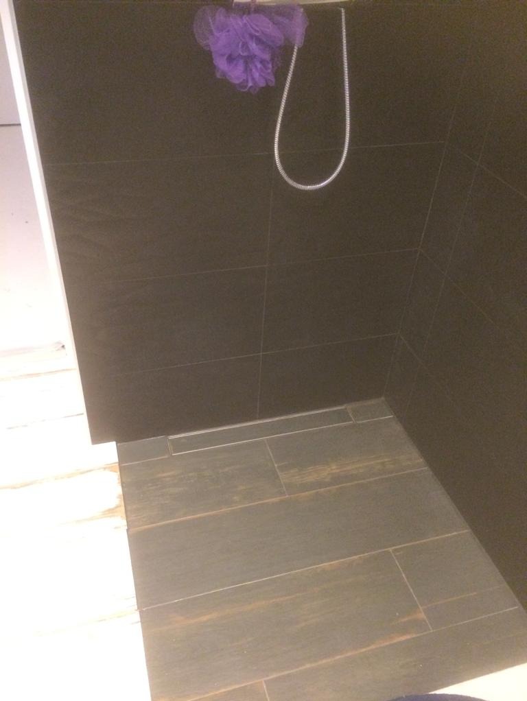 Ciemne płytki ścienne i podłogowe imitujące drewno w kabinie prysznicowej, widoczny odpływ liniowy i fioletowa myjka zawieszona na słuchawce prysznicowej.