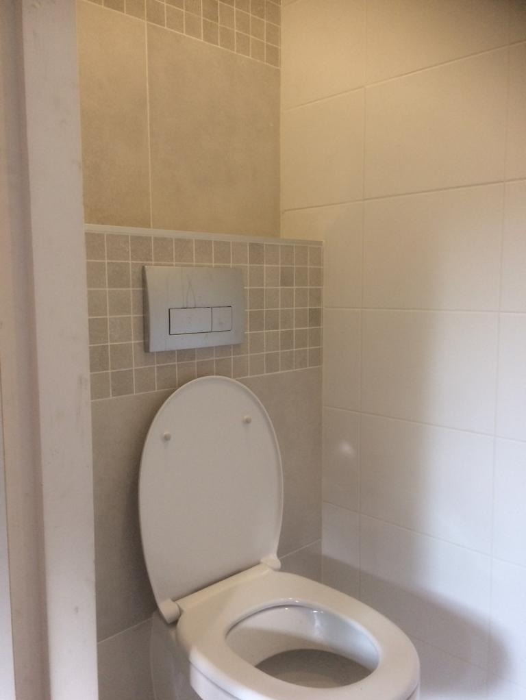 Biała toaleta z otwartą klapą w łazience z beżowymi płytkami, widoczny przycisk spłuczki na ścianie i fragment ościeżnicy drzwiowej po lewej stronie.