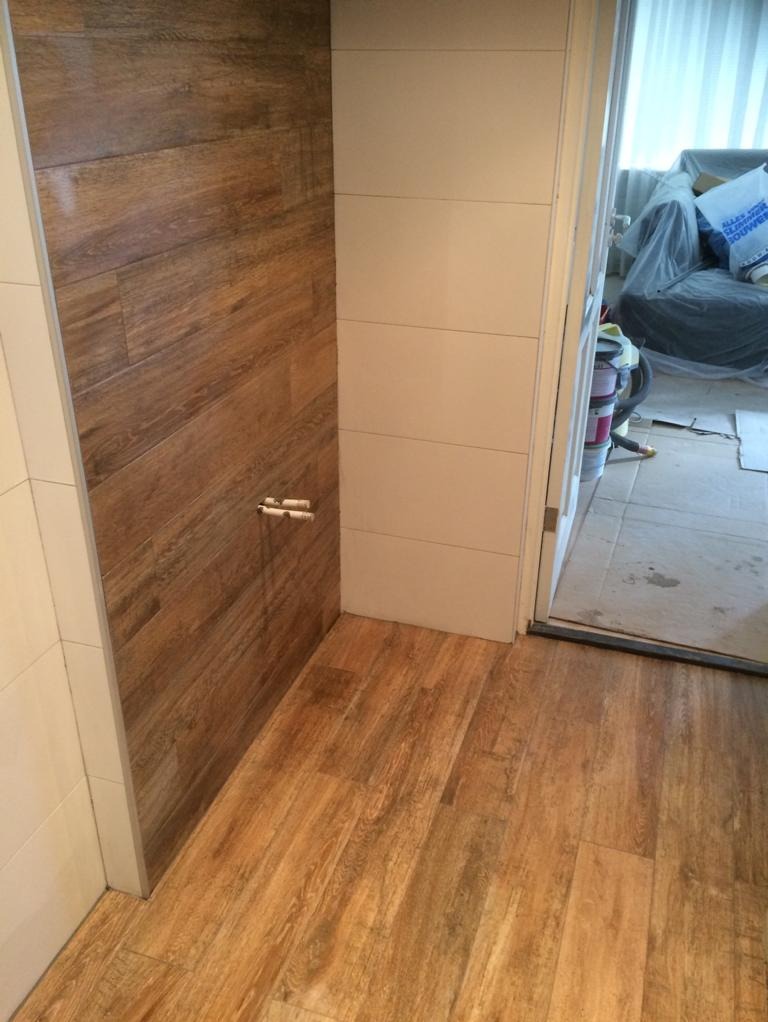 Wykończona ściana prysznicowa z imitacją drewna, jasna podłoga z paneli, fragment białej ściany z poziomymi podziałami, widoczne rury instalacji wodnej, w tle pomieszczenie w trakcie prac...