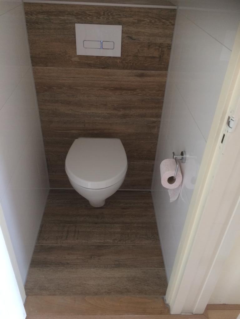 Nowoczesna toaleta w małej łazience, z drewnopodobnymi panelami na podłodze i ścianie za muszlą, białe płytki na ścianach bocznych, biała muszla klozetowa i przycisk spłuczki, uchwyt na papier...