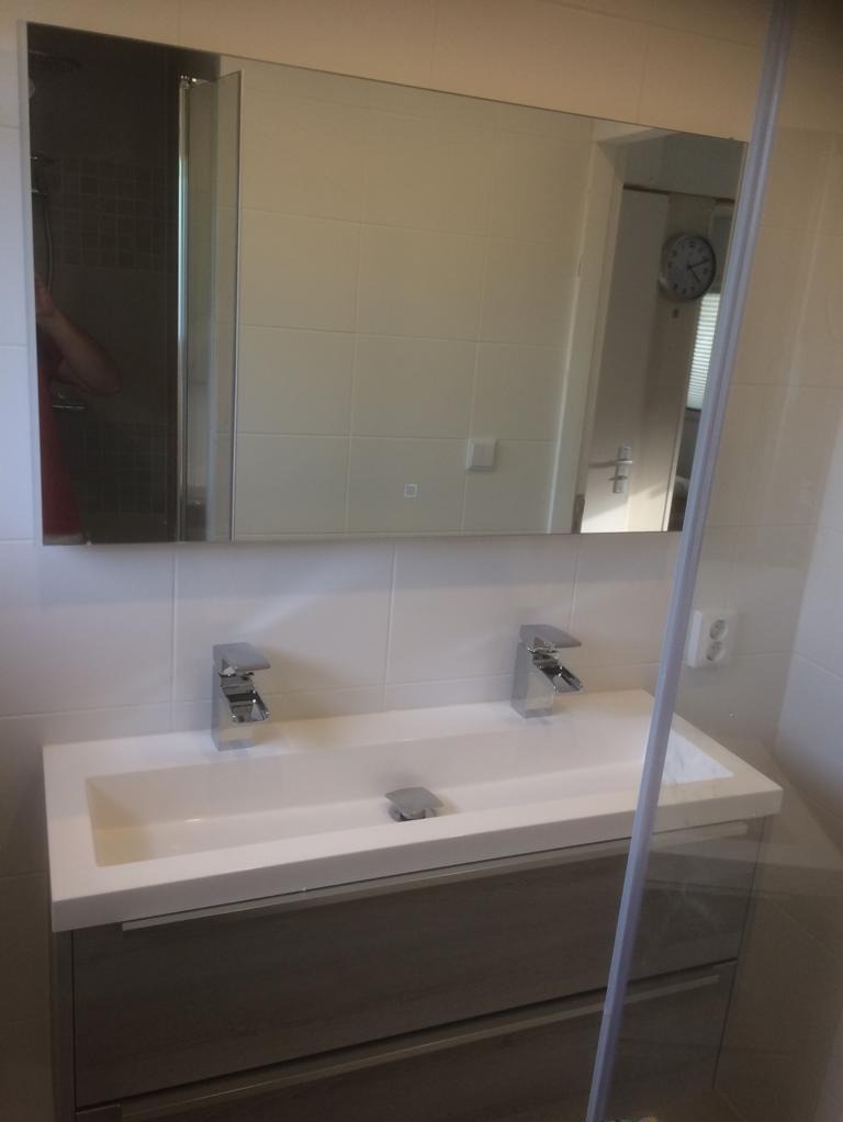 Nowoczesna umywalka nablatowa z dwiema chromowanymi bateriami, zamontowana na szafce z szufladami w odcieniach szarości, z dużym lustrem nad nią, odbijającym fragment kabiny prysznicowej i zegar...