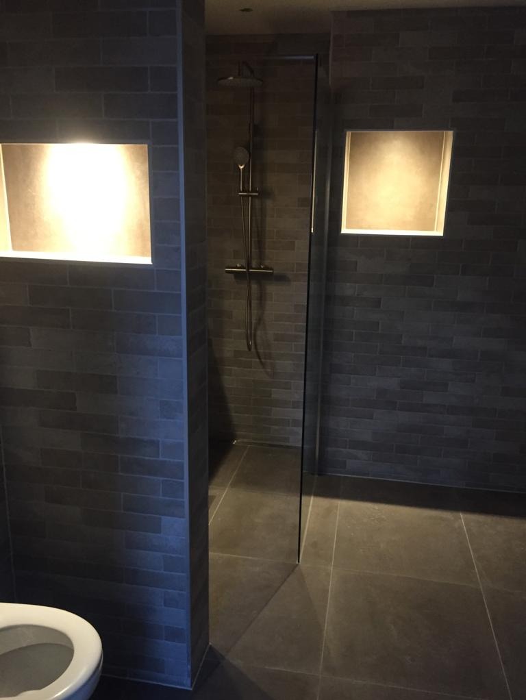 Nowoczesna łazienka z szarymi płytkami imitującymi cegłę, podświetlanymi wnękami i kabiną prysznicową typu walk-in; widoczny fragment toalety.