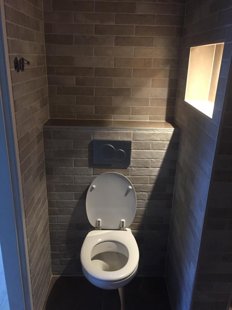 Kompaktowa toaleta z podtynkową spłuczką i wnęką oświetleniową w ścianie wyłożonej cegiełkowymi płytkami w odcieniach szarości i brązu.