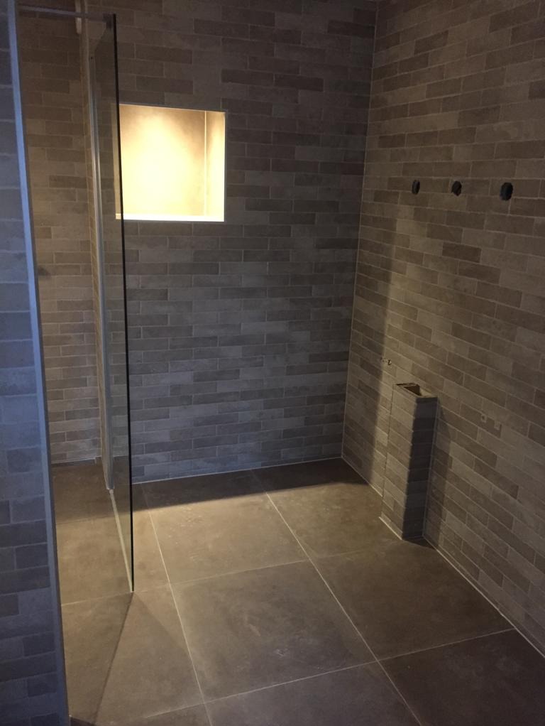 Nowoczesna łazienka z szarymi cegiełkami na ścianach, ciemnymi płytkami podłogowymi i wnęką oświetloną od wewnątrz, widoczna szklana kabina prysznicowa.