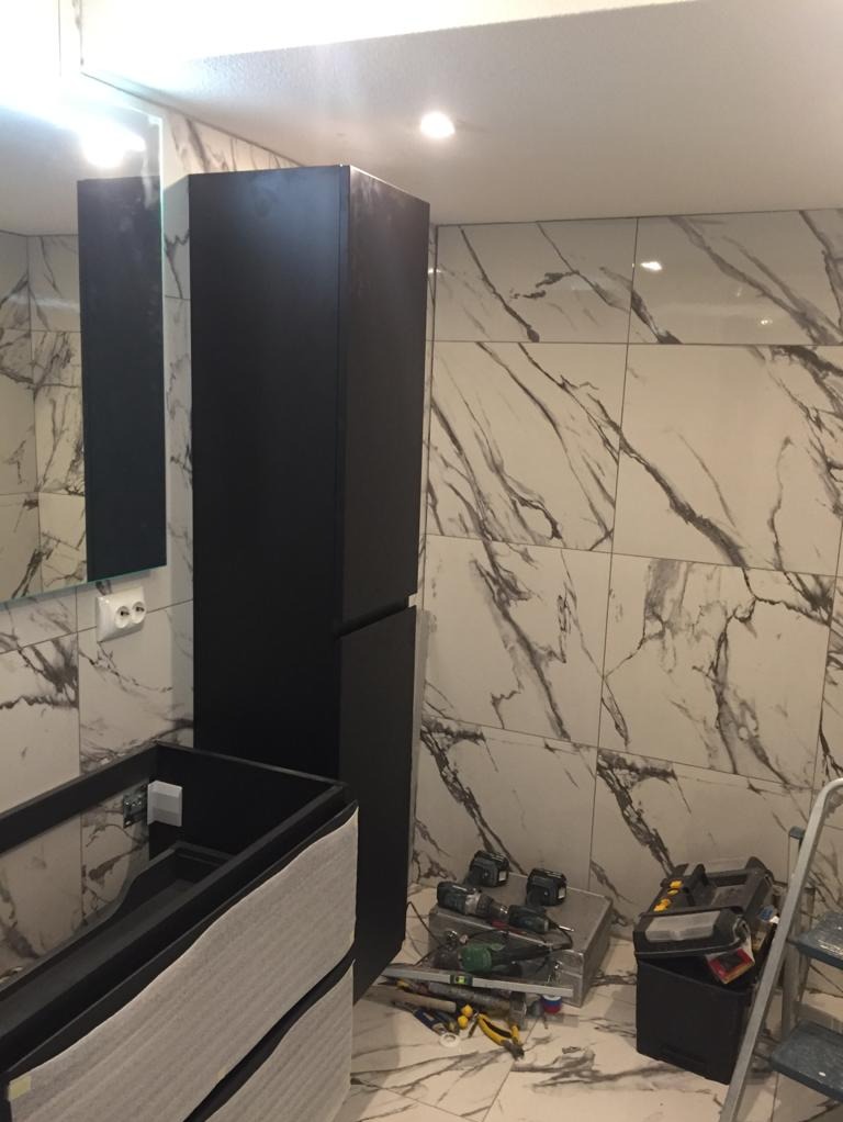 Montaż czarnej szafki łazienkowej z umywalką na tle marmurowych płytek, widoczne narzędzia i drabina.