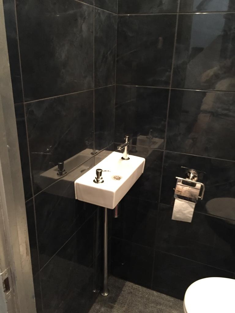 Kompaktowa umywalka z chromowanymi bateriami zamontowana na pojedynczej nodze, na tle czarnych, marmurowych płytek w małej łazience.