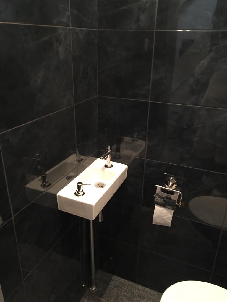 Wąska, prostokątna umywalka na chromowanej nodze w małej łazience wyłożonej czarnymi, marmurkowymi płytkami; widoczny fragment toalety i uchwyt na papier toaletowy.