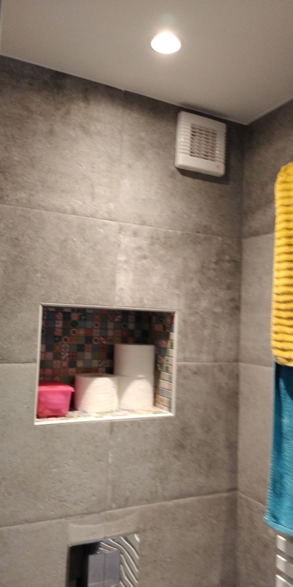 Szara łazienka z wnęką wyłożoną mozaiką, w której stoją rolki papieru toaletowego i różowy pojemnik, widoczny fragment grzejnika drabinkowego i wentylator wyciągowy.