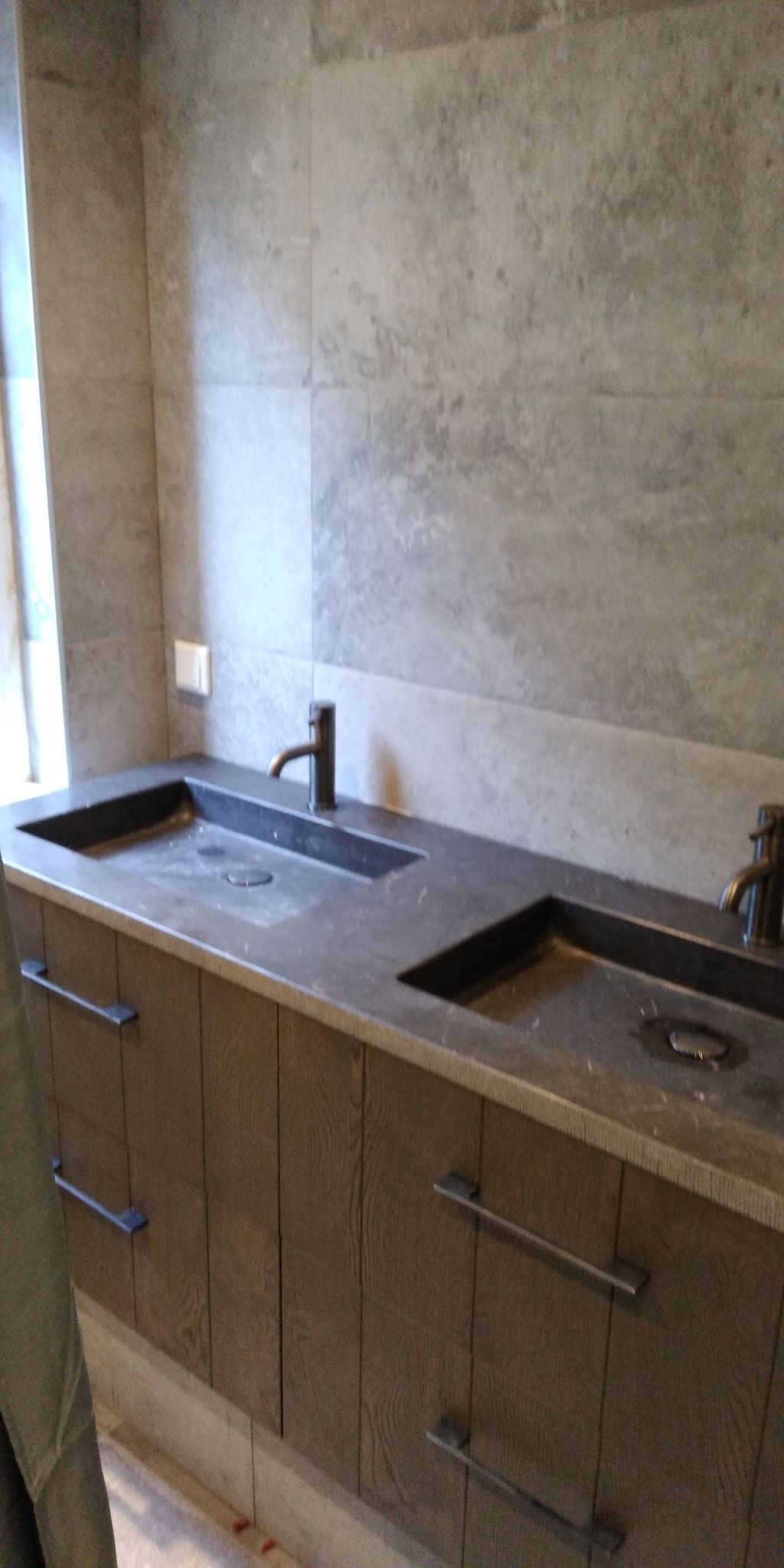 Dwie umywalki z ciemnego kamienia z brązowymi bateriami na drewnianej szafce z szufladami w łazience wykończonej szarymi płytkami.