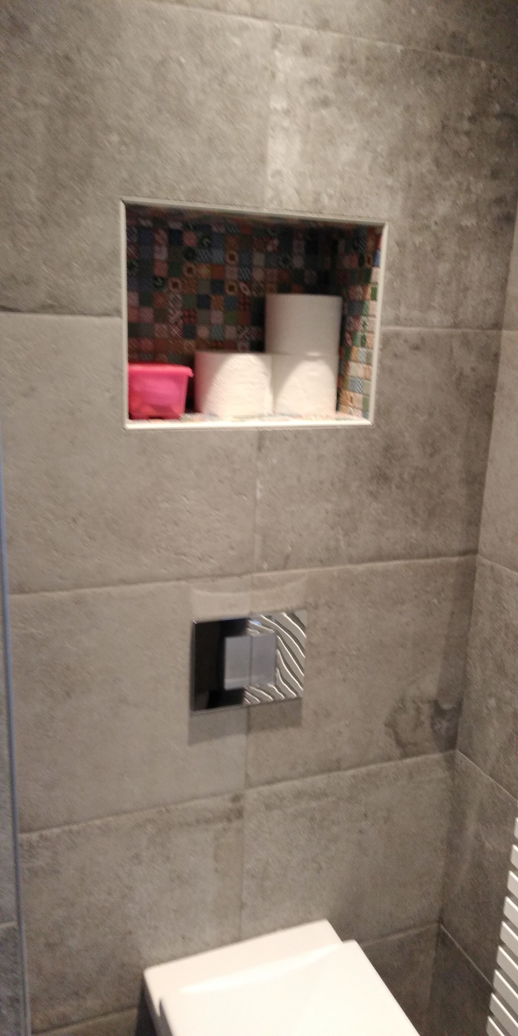 Szara łazienka z wnęką wyłożoną mozaiką, w której znajdują się rolki papieru toaletowego i różowy pojemnik. Widoczny fragment toalety i chromowany przycisk spłuczki.